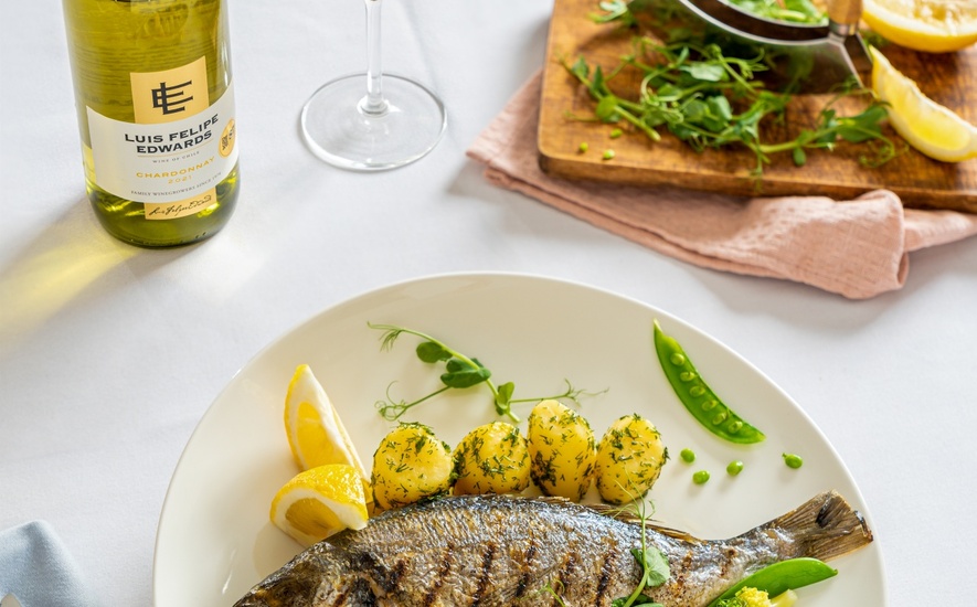 Grillowana dorada w całości z koperkowymi ziemniakami i sezonowymi warzywami, podana z kieliszkiem i butelką Chardonnay.