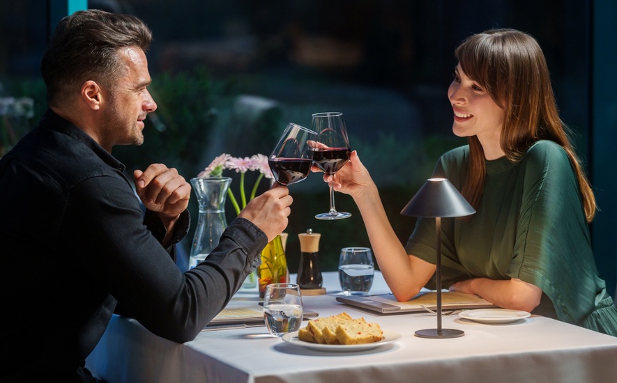 Para wznosi toast czerwonym winem przy przytulnie oświetlonym stole w restauracyjnej części hotelu, chleb, woda, kwiaty, lampka