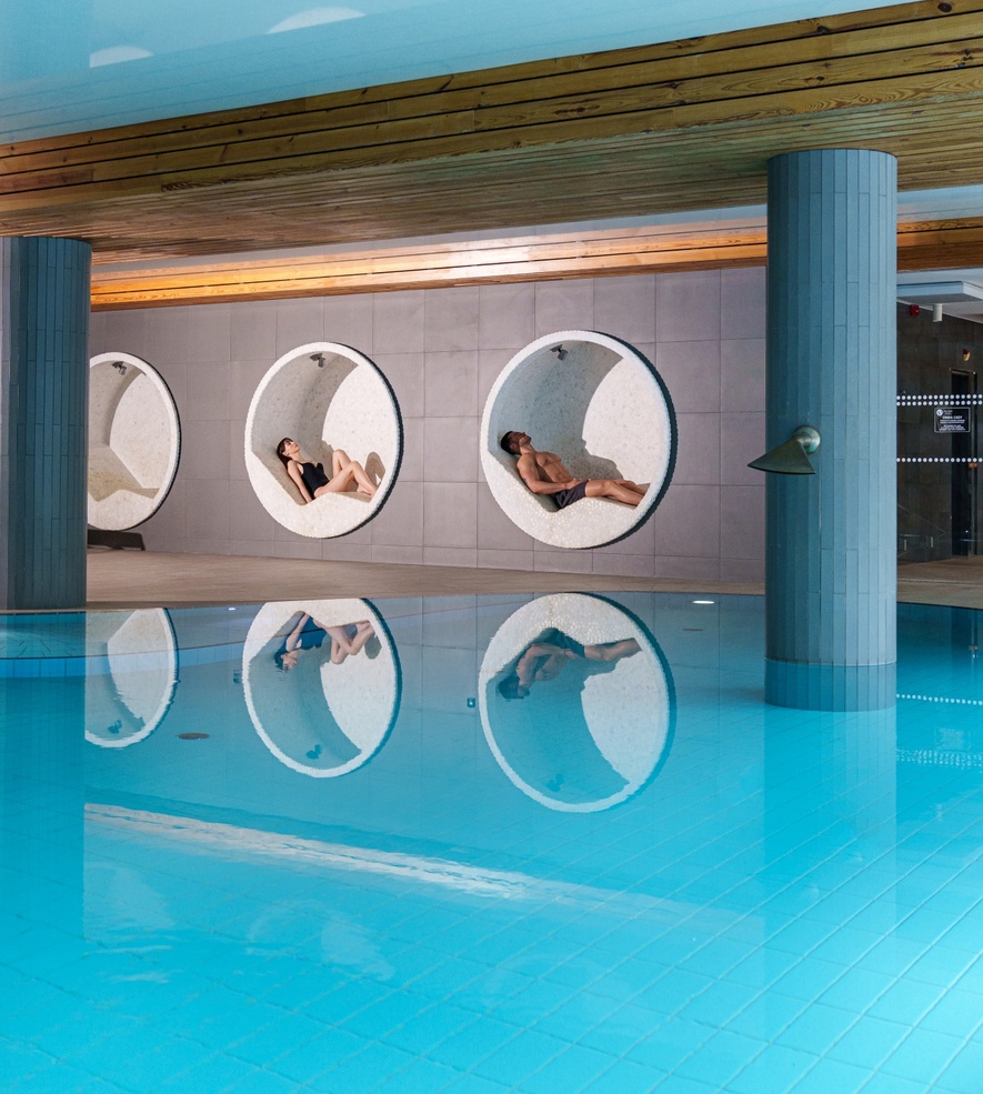 Indoor hotel pool with turquoise tiles, wooden ceiling and circular white lounge alcoves where two guests recline, mirrored in water.