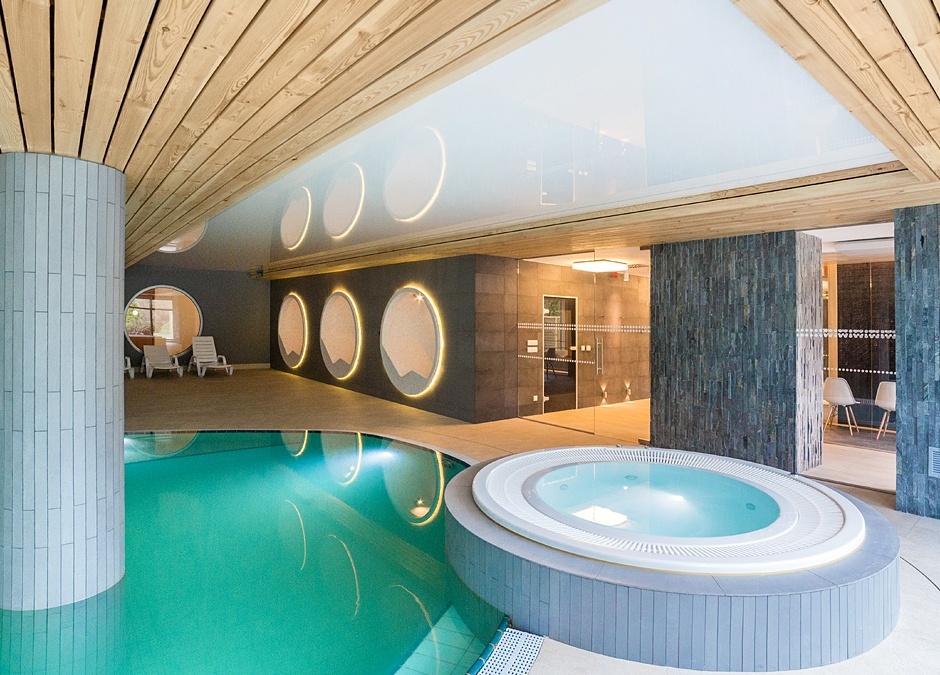 Spa wewnętrzne z turkusowym basenem, okrągłym jacuzzi, drewnianym sufitem, podświetlanymi oknami i leżakami.
