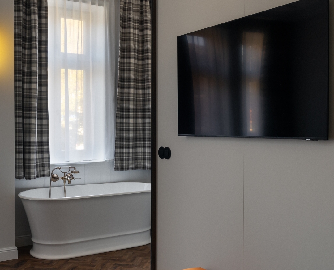 Apartament z wolnostojącą wanną przy oknie, zasłonami w kratę, telewizorem na ścianie i pomarańczowym pufem