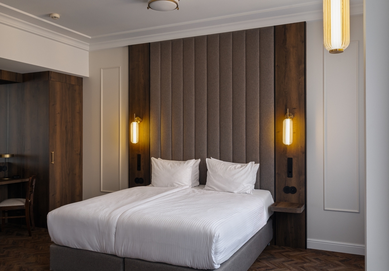 Nowoczesny pokój z dwoma łóżkami złączonymi w podwójne, wysokim tapicerowanym zagłówkiem, ciepłymi lampami, drewniana szafa i biurko.