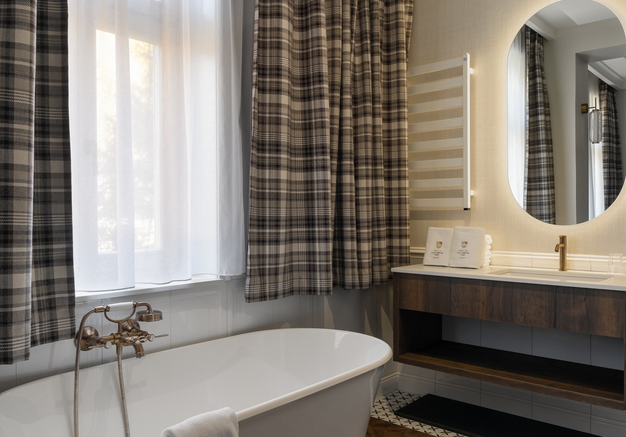 Wolnostojąca wanna z mosiężną baterią przy oknie z firanką i zasłoną w kratę, podświetlane lustro i szafka z ręcznikami.