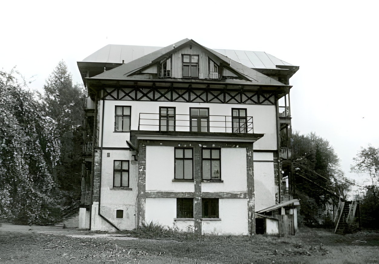Historyczna willa z drewnianą konstrukcją, balkonem i zewnętrznymi schodami, w zalesionym otoczeniu.
