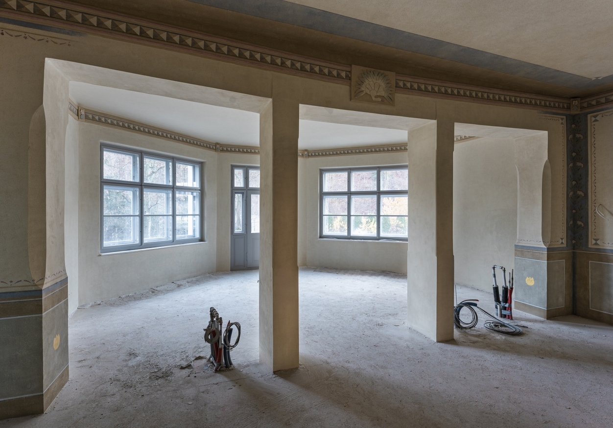 Przestronne zabytkowe pomieszczenie w remoncie z dekoracyjnym fryzem, arkadowymi filarami, dużymi oknami i odsłoniętymi przewodami na surowej podłodze