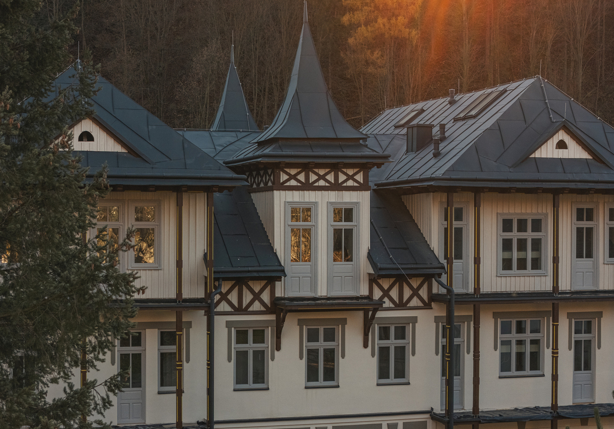 Fasada willi w stylu alpejskim z wieżyczkami, ciemnymi metalowymi dachami i białym drewnianym obiciem na tle zalesionego wzgórza o zachodzie słońca
