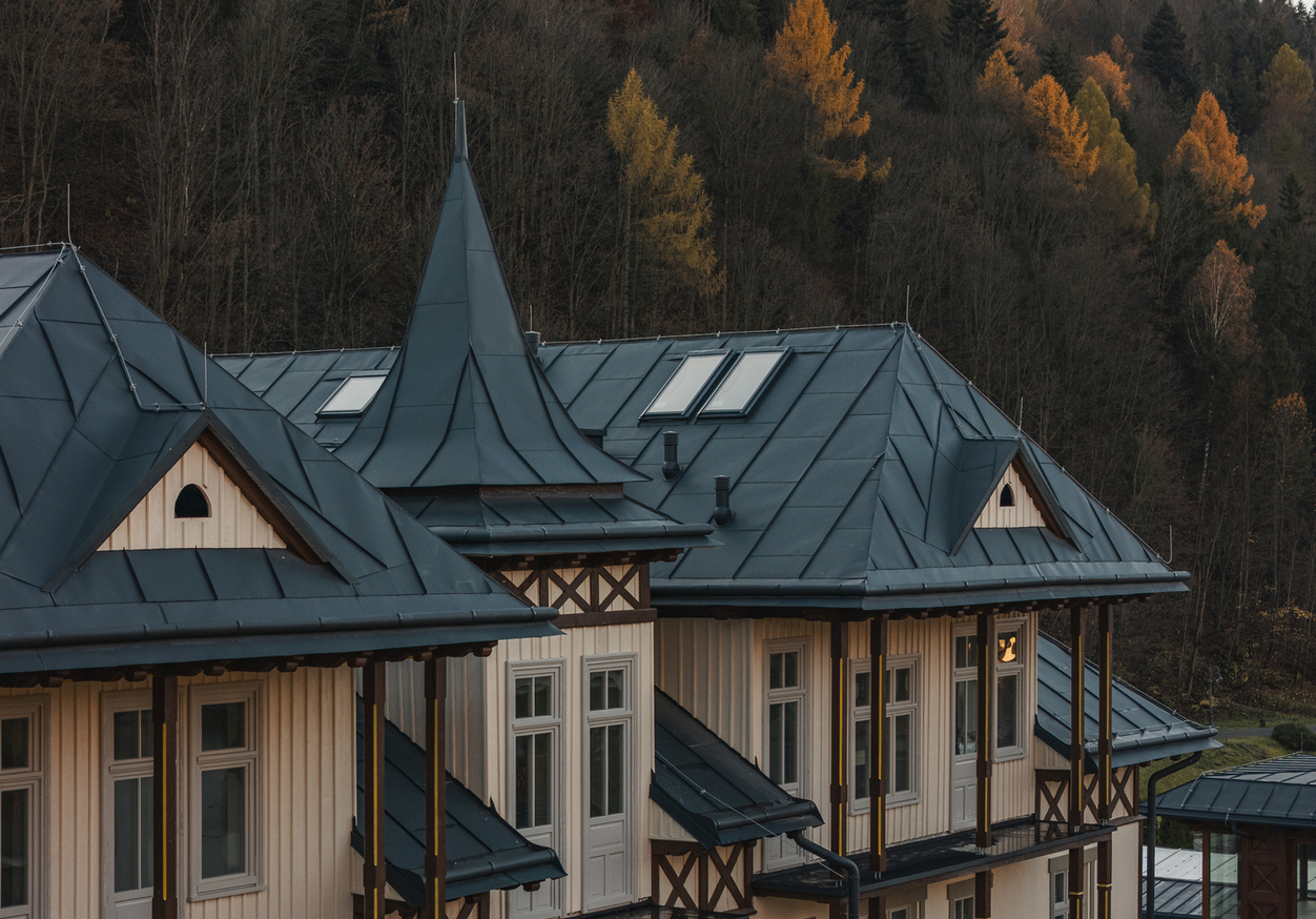 Fasada hotelu w alpejskim stylu z ciemnymi metalowymi dachami, wieżyczką, drewnianymi balkonami i oknami na tle jesiennego lasu.