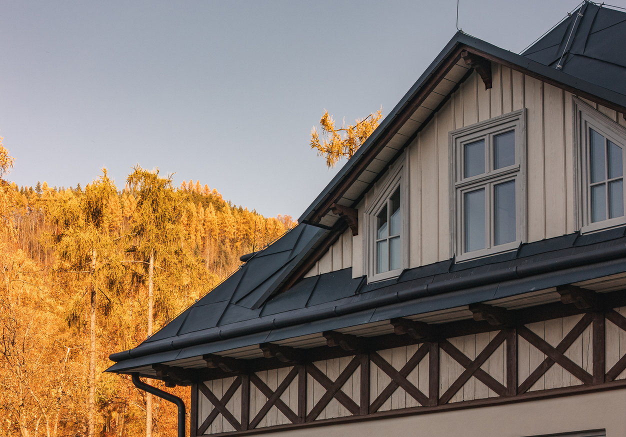 Szczyt górskiego pensjonatu z oknami mansardowymi, ciemnym metalowym dachem i drewnianymi belkami, złoty jesienny las w tle.