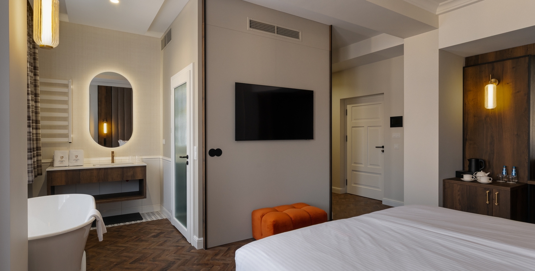 Nowoczesny pokój z wolnostojącą wanną, podświetlanym owalnym lustrem, telewizorem na ścianie, pomarańczowym pufem i minibarem z czajnikiem