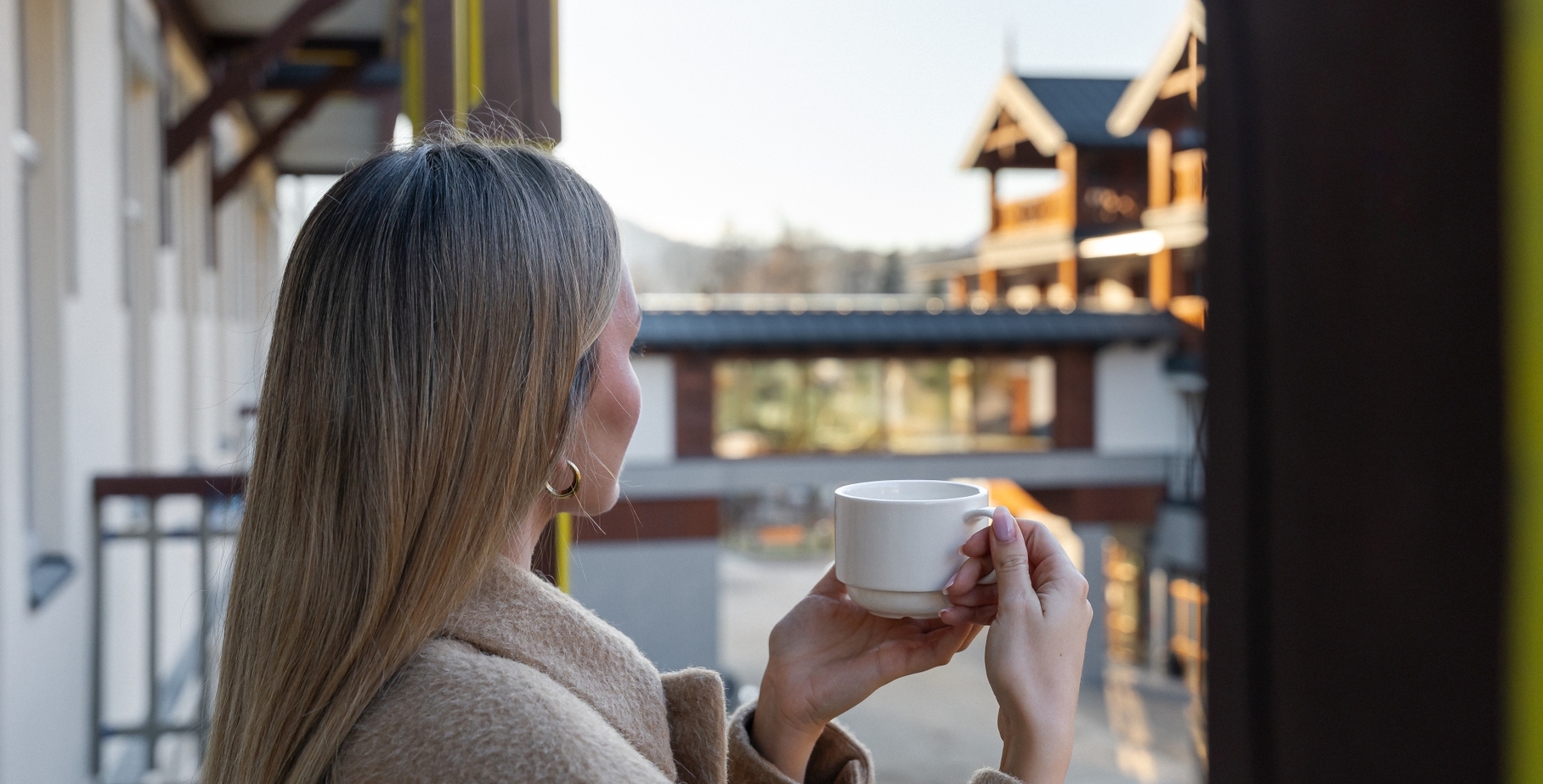 Gość na balkonie trzyma biały kubek, w ciepłym płaszczu, z widokiem na drewniane budynki hotelu i odległe góry