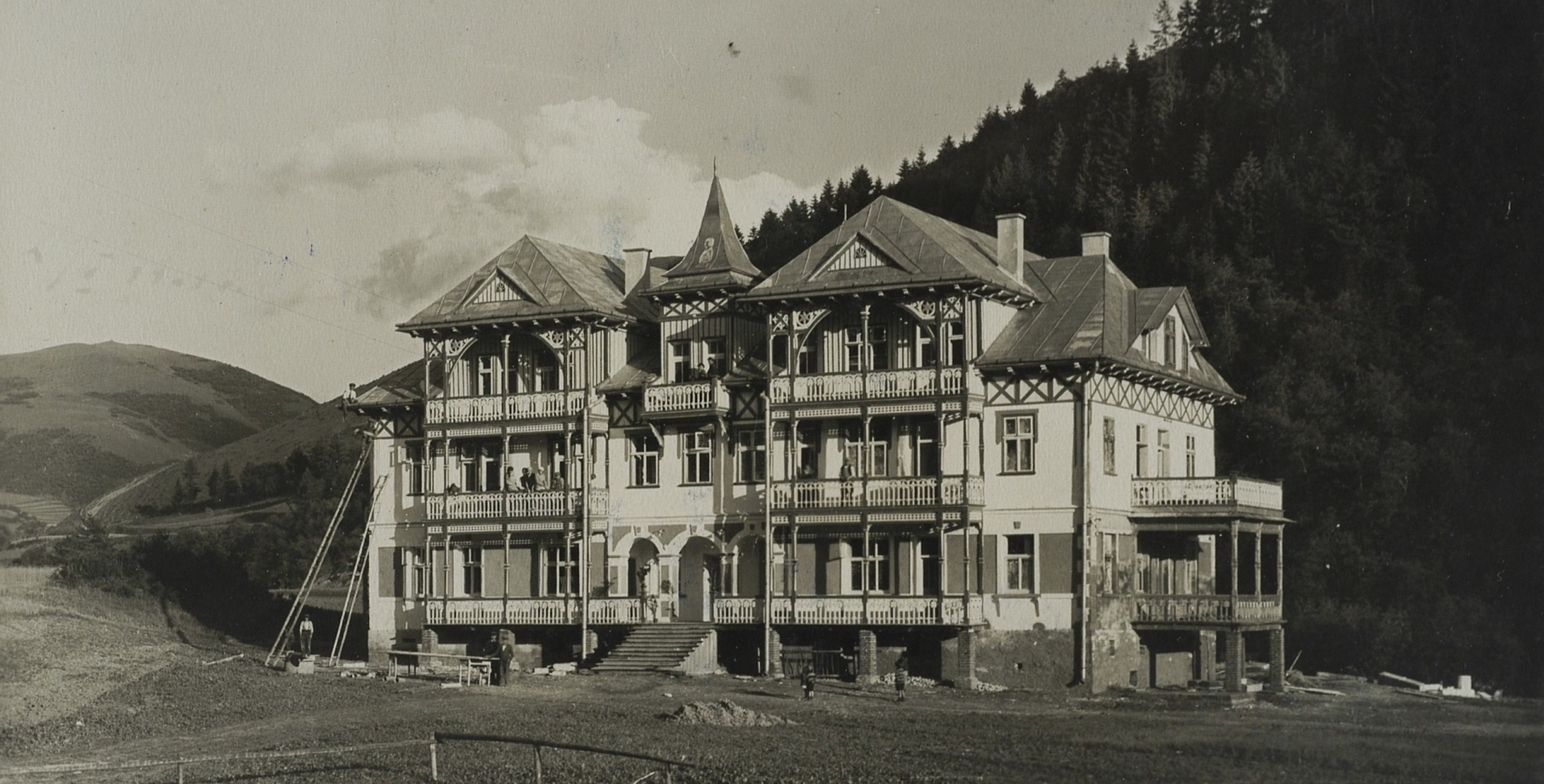 Zabytkowa górska willa z misternymi drewnianymi balkonami i tarasami, położona u stoku zalesionych wzgórz.