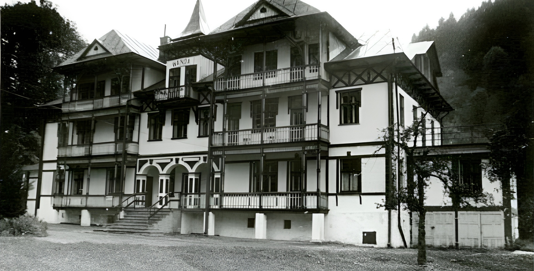 Zabytkowy górski hotel z drewnianymi balkonami i zadaszonymi werandami, wielopiętrowa elewacja na tle zalesionego stoku.