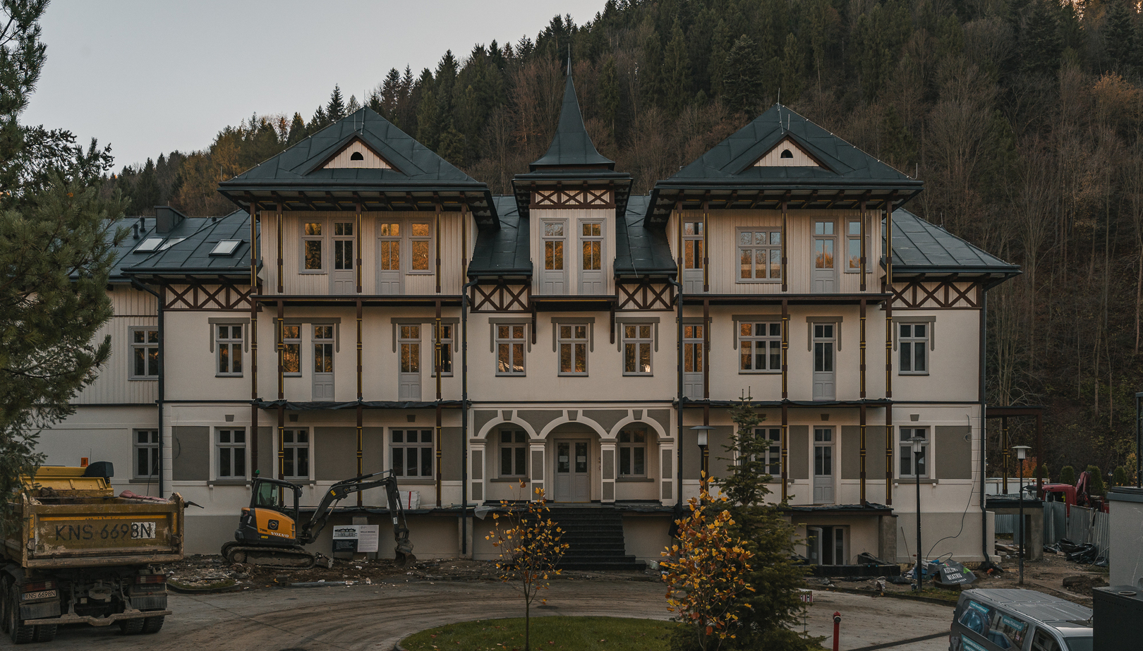 Odnowiona górska willa hotelowa z dekoracyjnymi balkonami i arkadowym wejściem, las w tle oraz widoczne maszyny budowlane.