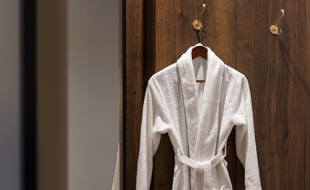 Miękki biały szlafrok z paskiem wiszący na mosiężnych haczykach na ciepłej drewnianej ścianie obok lustra i podłogi w jodełkę