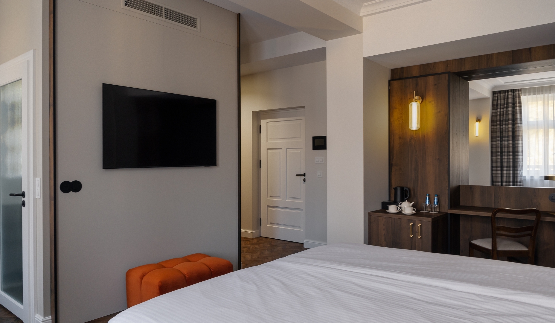 Nowoczesny pokój z łóżkiem, telewizorem na ścianie, pomarańczową pufą, drewnianym biurkiem z czajnikiem i butelkowaną wodą, ciepłe oświetlenie.