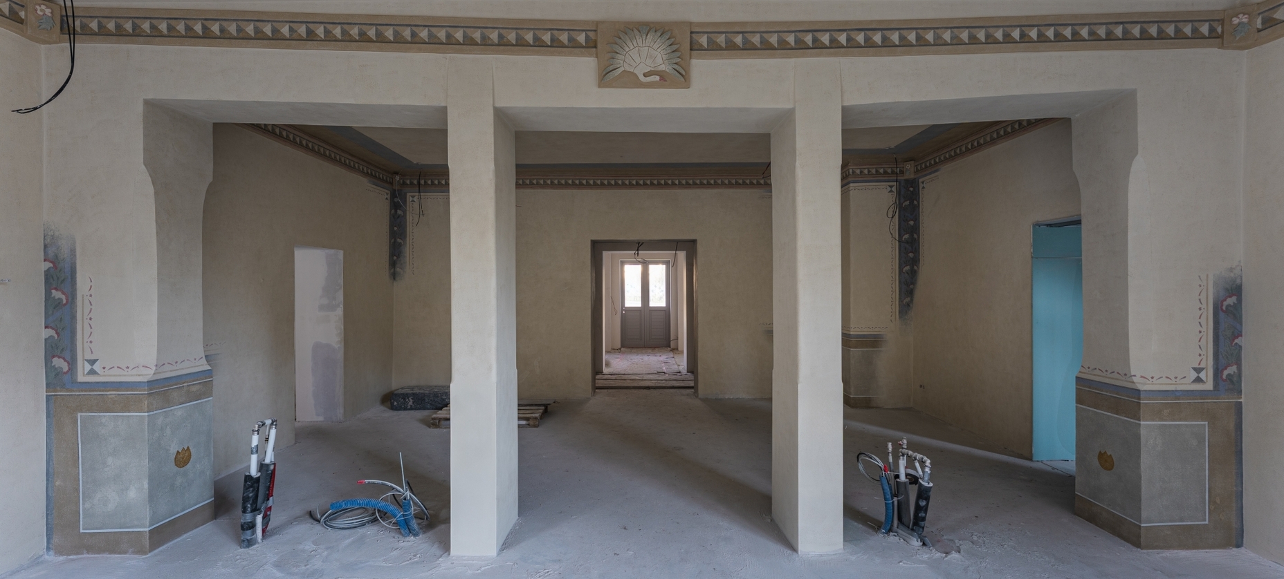 Historyczny pokój hotelowy w remoncie z ozdobnym gzymsem i freskami, trzema kolumnami i łukowymi niszami, odsłonięte rury.