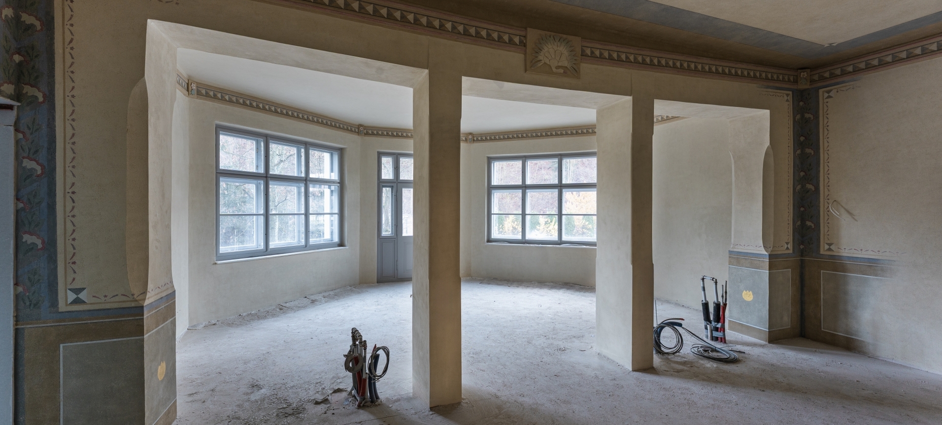 Przestronne zabytkowe pomieszczenie w remoncie z dekoracyjnym fryzem, arkadowymi filarami, dużymi oknami i odsłoniętymi przewodami na surowej podłodze