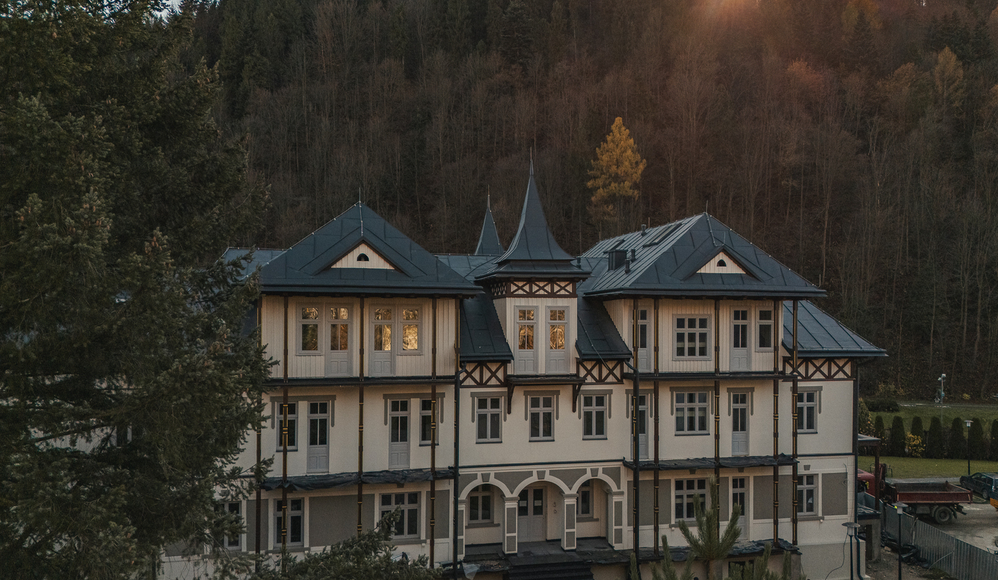 Zabytkowy górski pensjonat z trzema kondygnacjami, wieżyczką, łukowym wejściem i dużymi oknami na tle zalesionego stoku o zachodzie słońca.