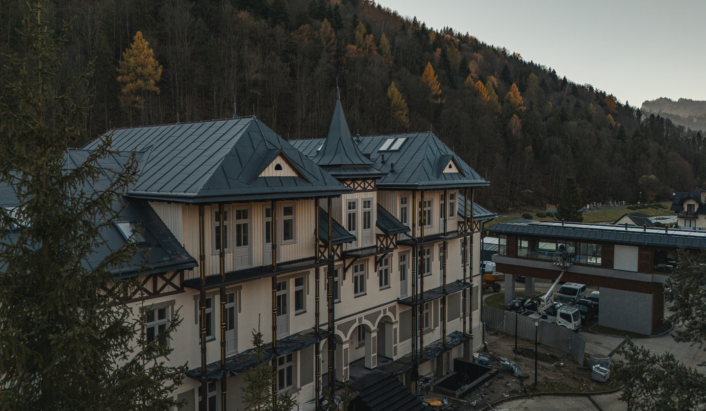 Górski hotel w stylu alpejskim z wieżyczką i balkonami, blisko jesiennego lasu; przeszklone przejście i prace remontowe.