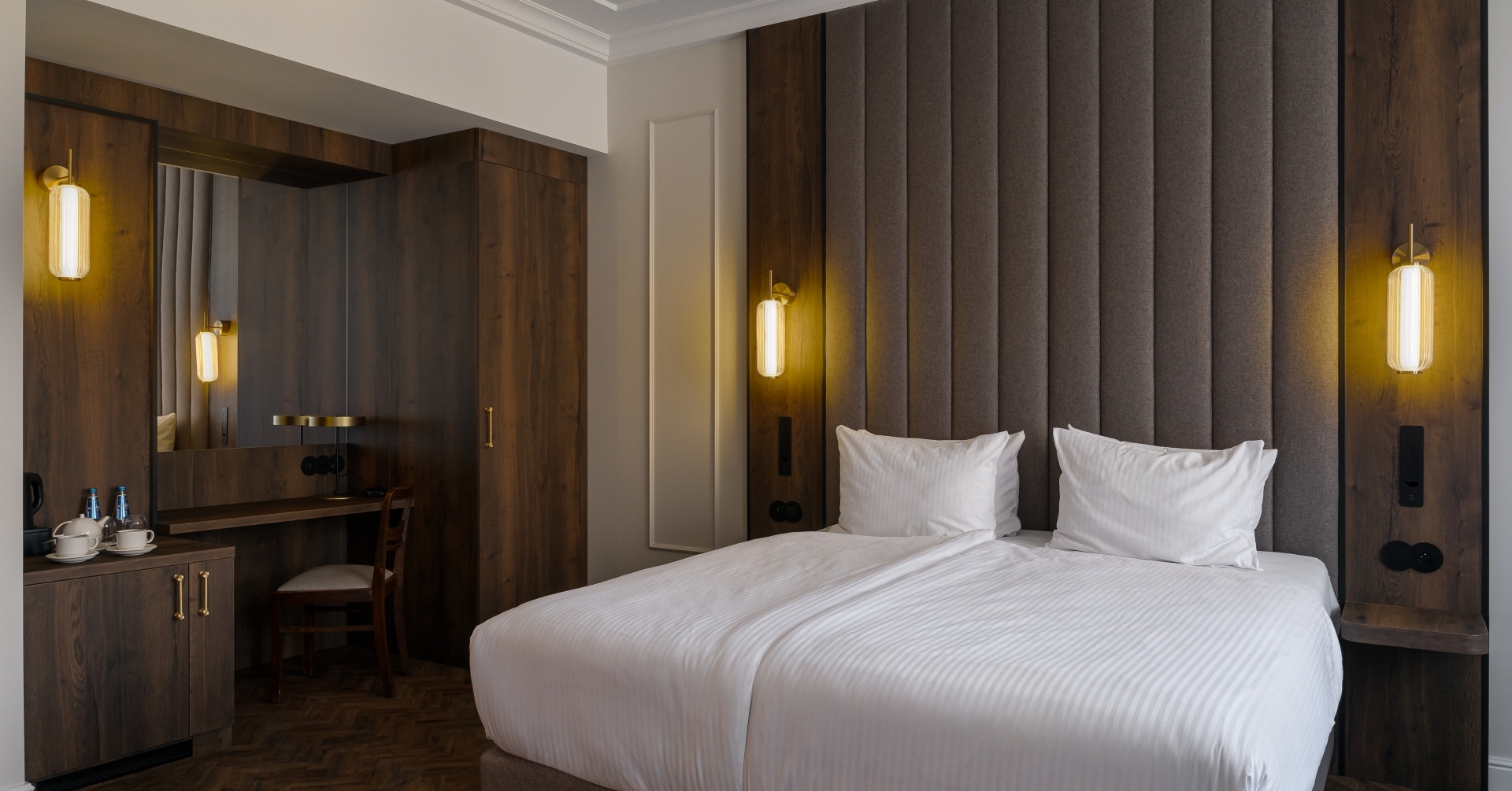 Nowoczesny pokój z dwoma łóżkami, wysokim szarym wezgłowiem, mosiężnymi lampami, drewnianą zabudową, biurkiem i czajnikiem.