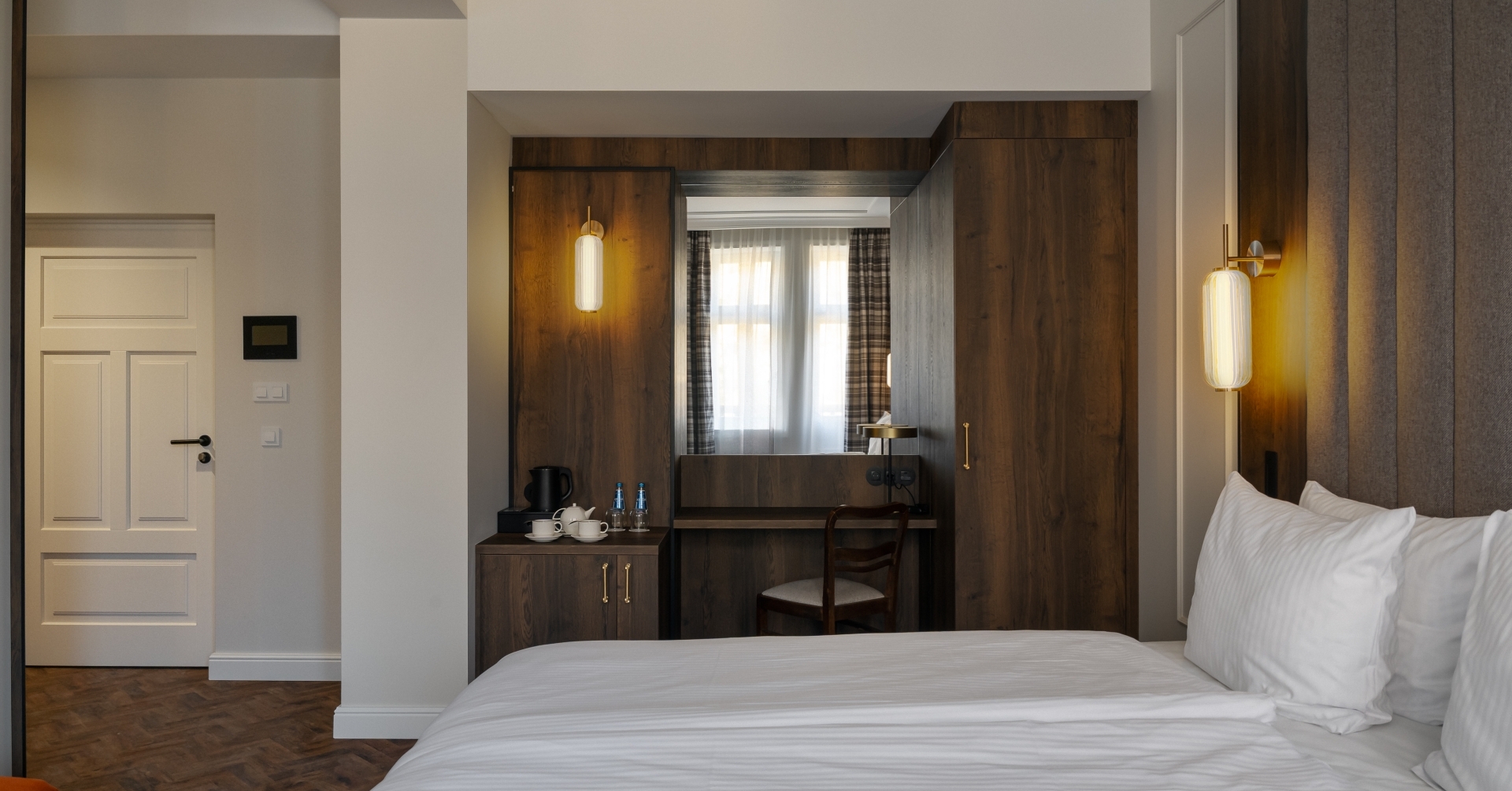 Przytulny pokój dwuosobowy z białą pościelą, tapicerowanym zagłówkiem, ciepłymi kinkietami, drewnianą szafą i czajnikiem.