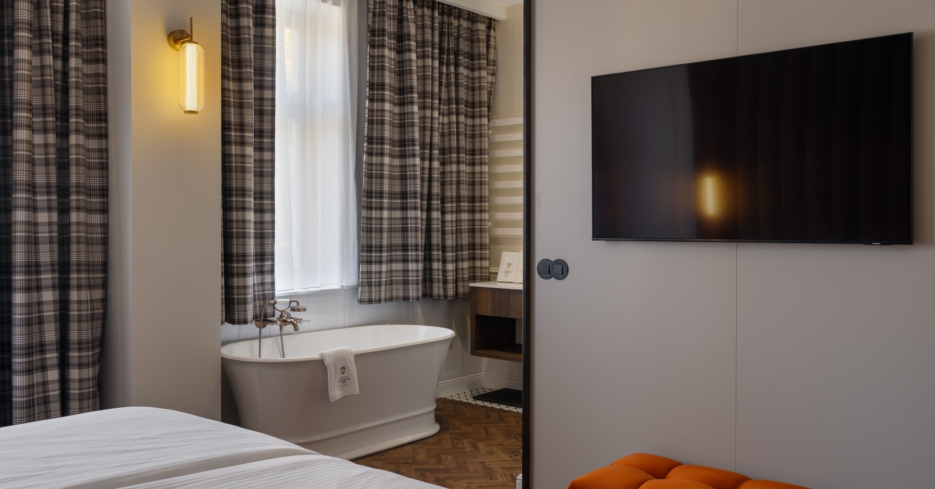 Wolnostojąca biała wanna przy oknie z zasłonami w kratę, ciepły kinkiet, telewizor na ścianie i pomarańczowy puf.