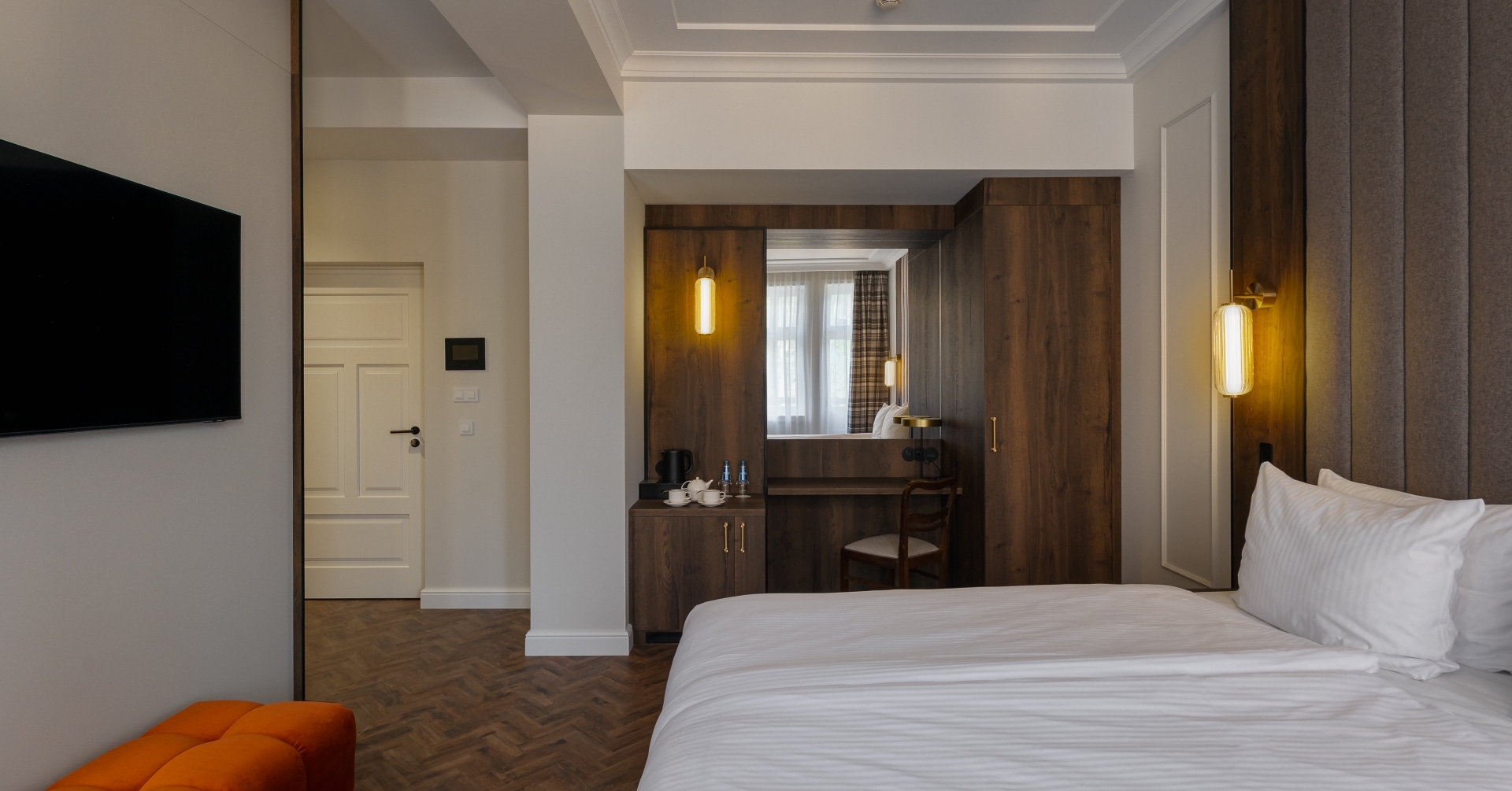 Nowoczesny pokój z łóżkiem king, tapicerowanym wezgłowiem, ciepłymi kinkietami, drewnianą szafą, biurkiem i telewizorem.