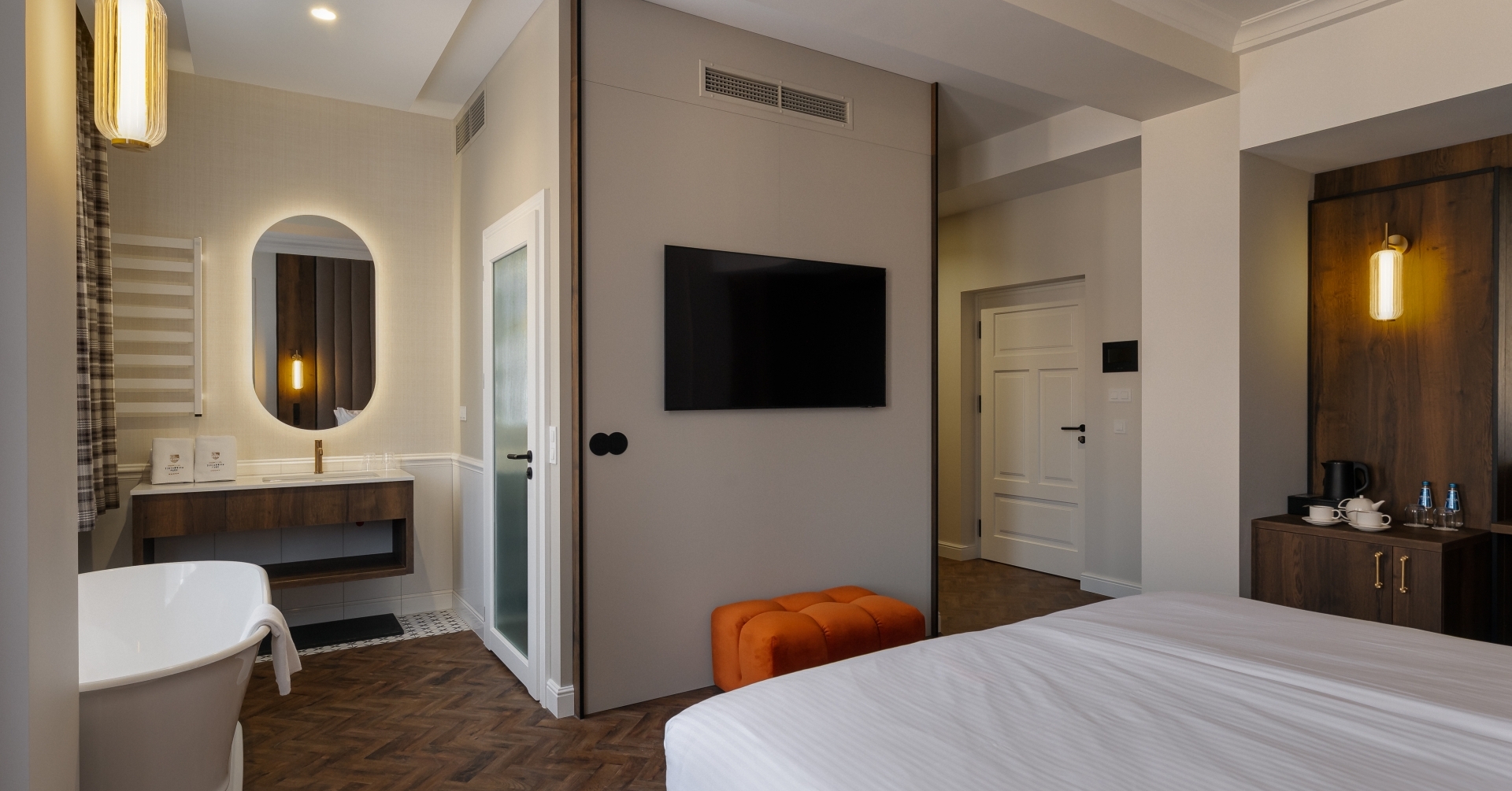 Nowoczesny pokój z wolnostojącą wanną, podświetlanym owalnym lustrem, telewizorem na ścianie, pomarańczowym pufem i minibarem z czajnikiem