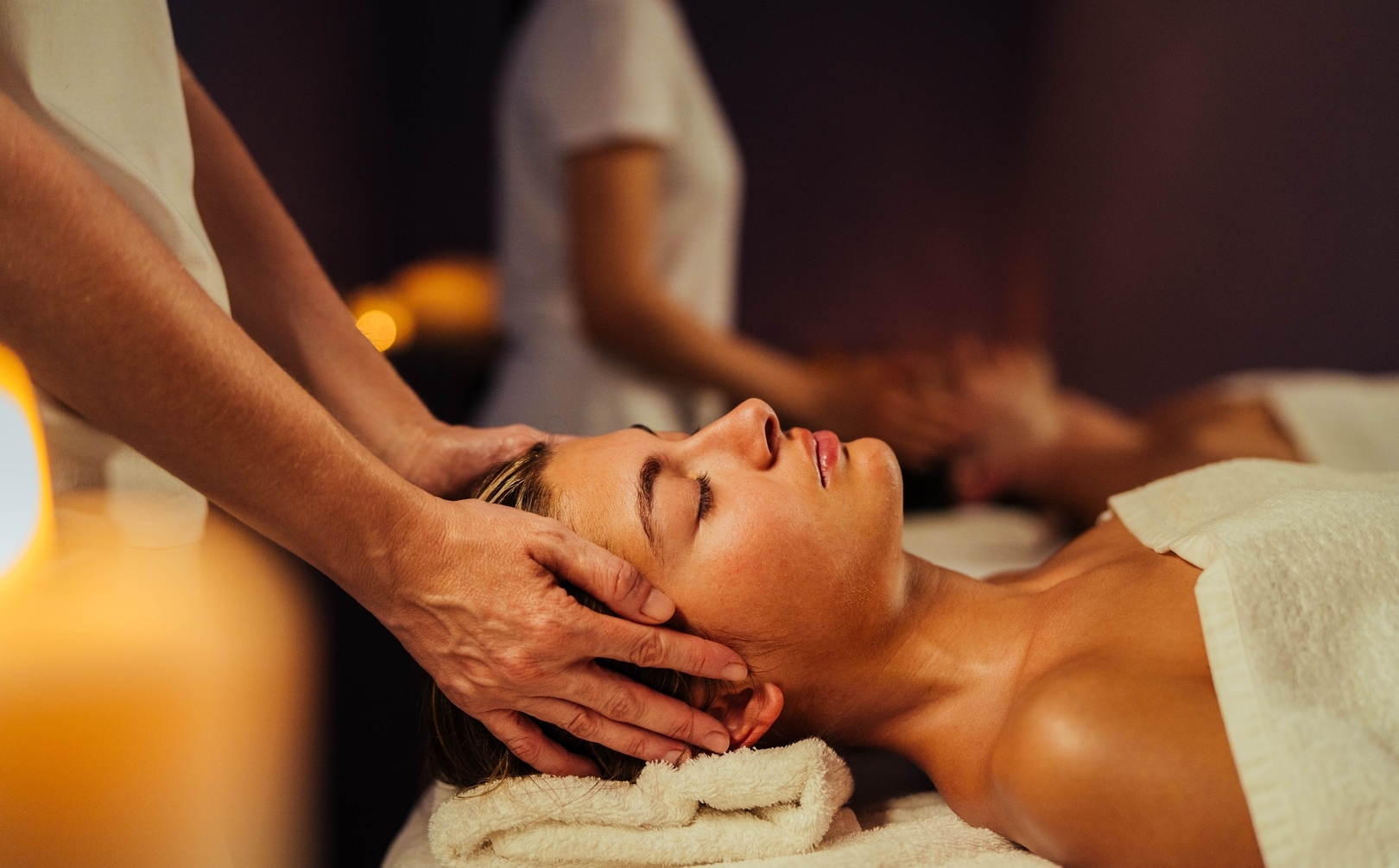 Kobieta otrzymuje relaksacyjny masaż głowy na stole spa, z ręcznikiem pod karkiem i ciepłym światłem świec.
