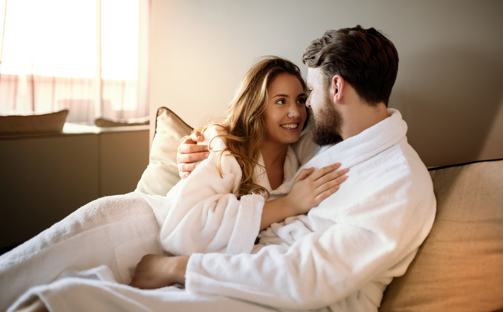 Para w białych szlafrokach przytulona na sofie w słonecznym apartamencie, kameralna, relaksująca atmosfera