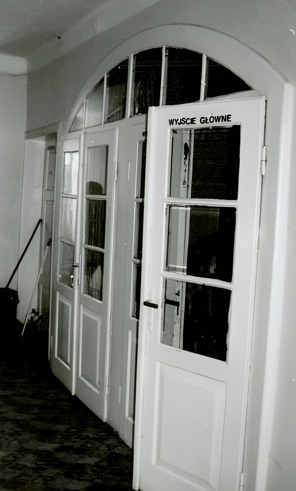 Białe przeszklone drzwi z łukowym nadprożem i napisem 'WYJŚCIE GŁÓWNE', kamienna posadzka i sprzęt porządkowy
