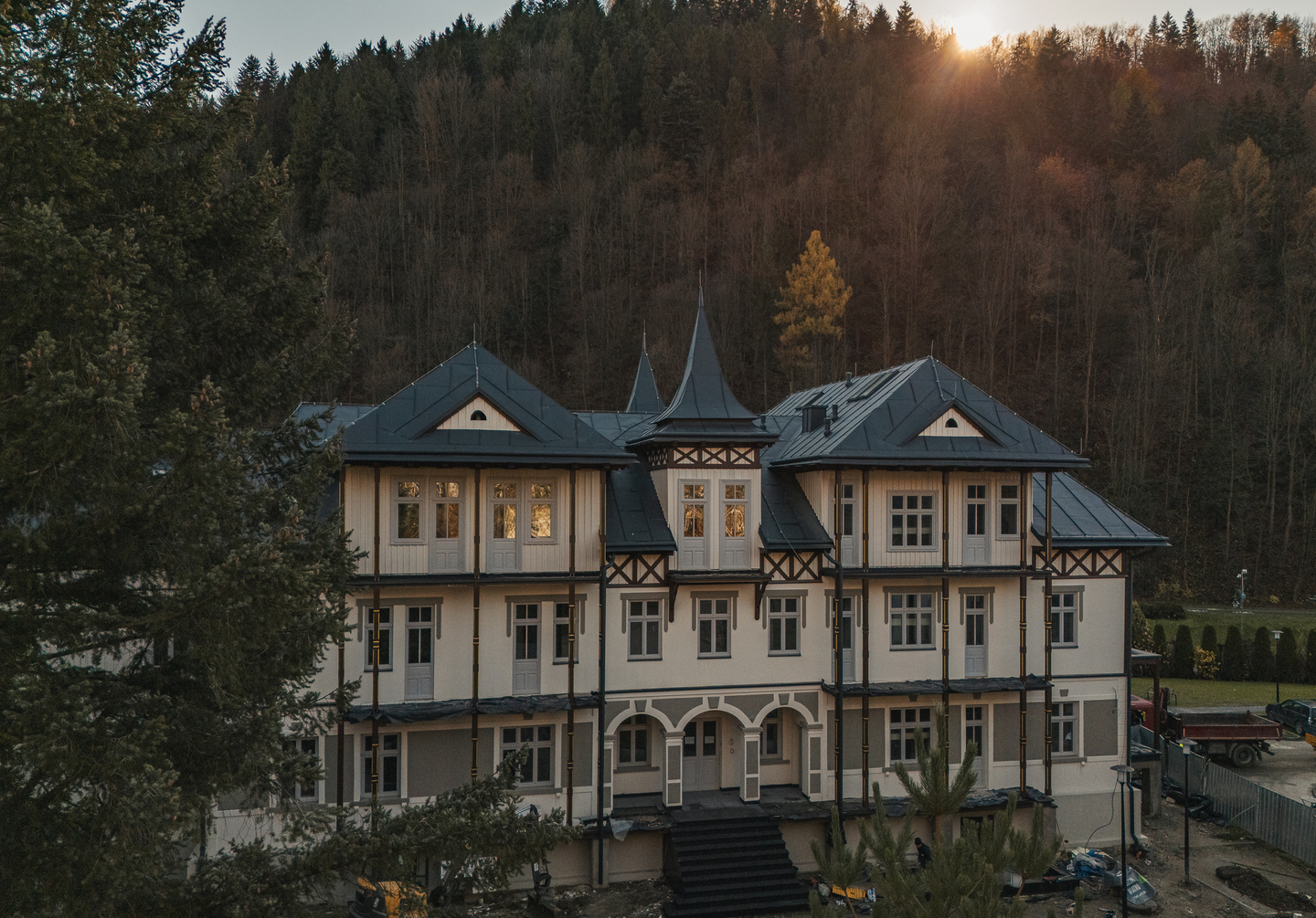 Zabytkowy górski pensjonat z trzema kondygnacjami, wieżyczką, łukowym wejściem i dużymi oknami na tle zalesionego stoku o zachodzie słońca.