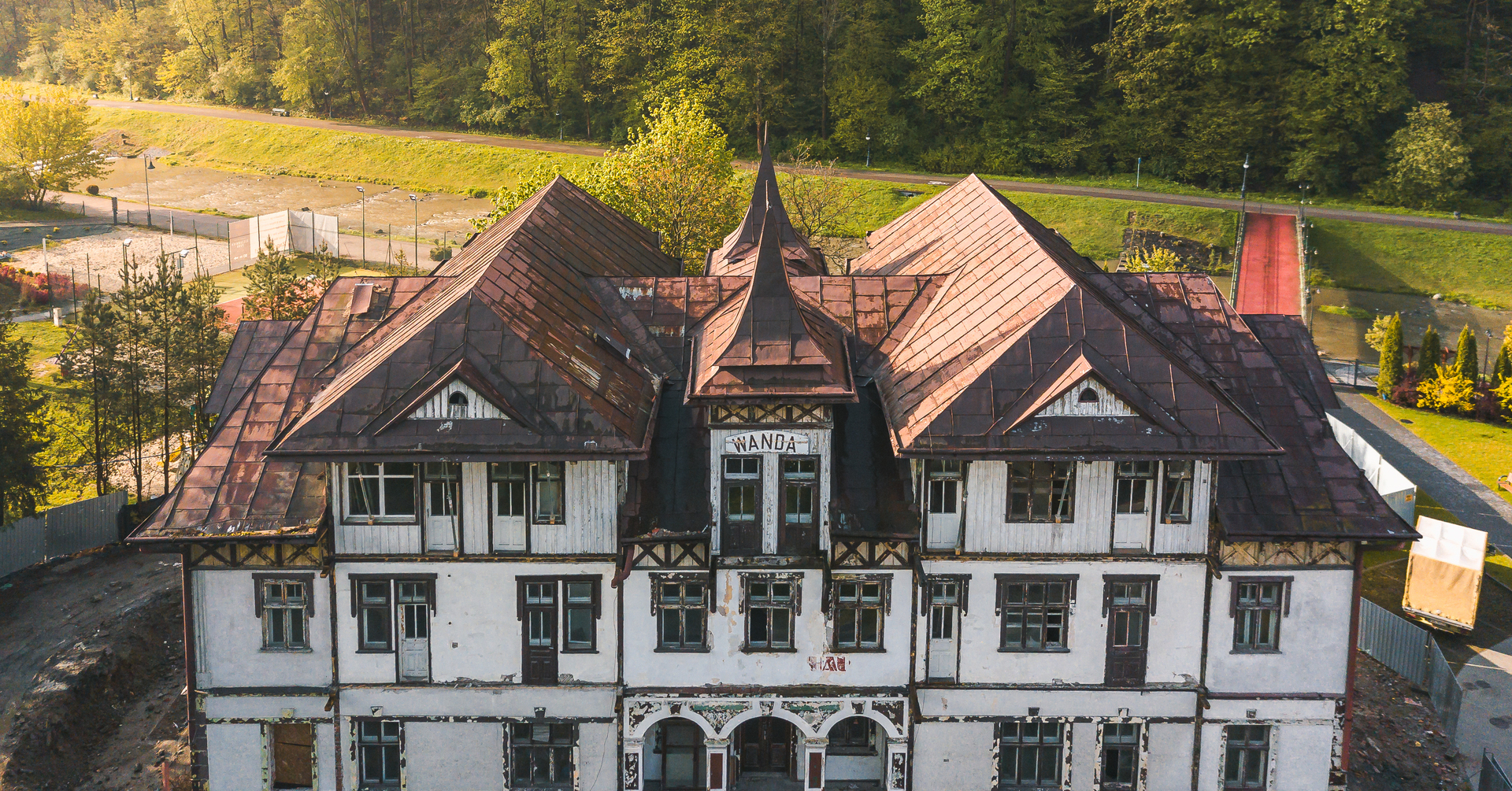 Zabytkowy górski hotel z łuszczącą się białą elewacją, zardzewiałymi spadzistymi dachami i wieżyczką, nad rzeką w leśnym otoczeniu.