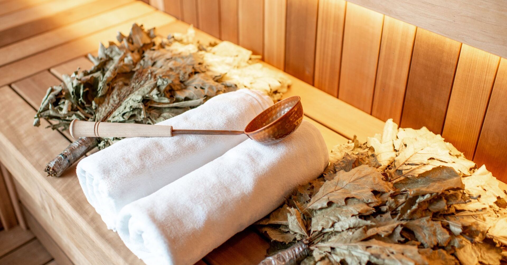 Ławka w saunie z drewnianymi panelami, zwiniętymi białymi ręcznikami, miedzianą chochelką i suszonymi wiązkami liści