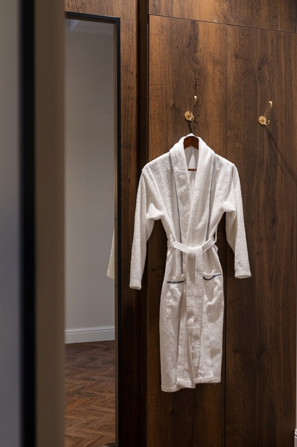 Miękki biały szlafrok z paskiem wiszący na mosiężnych haczykach na ciepłej drewnianej ścianie obok lustra i podłogi w jodełkę