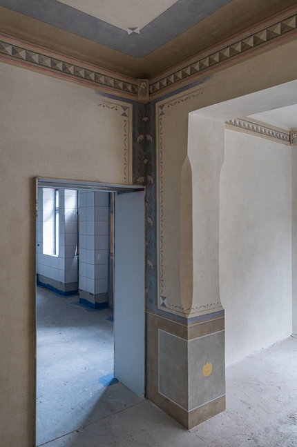 Historyczny korytarz z malowaną dekoracją i łukowym przejściem prowadzącym do jasnego, wykafelkowanego pomieszczenia