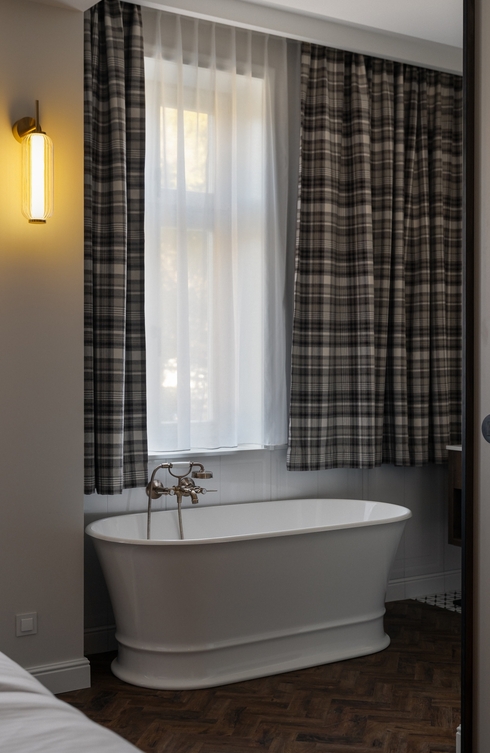 Wolnostojąca biała wanna z retro mosiężną baterią pod przezroczystą zasłoną, kraciaste firany, ciepły kinkiet i drewniana podłoga.