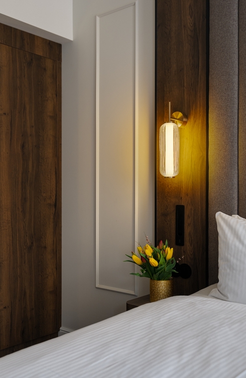 Ciepły kinkiet nad drewnianym zagłówkiem, śnieżnobiała pościel i złoty wazon z żółtymi tulipanami na stoliku.