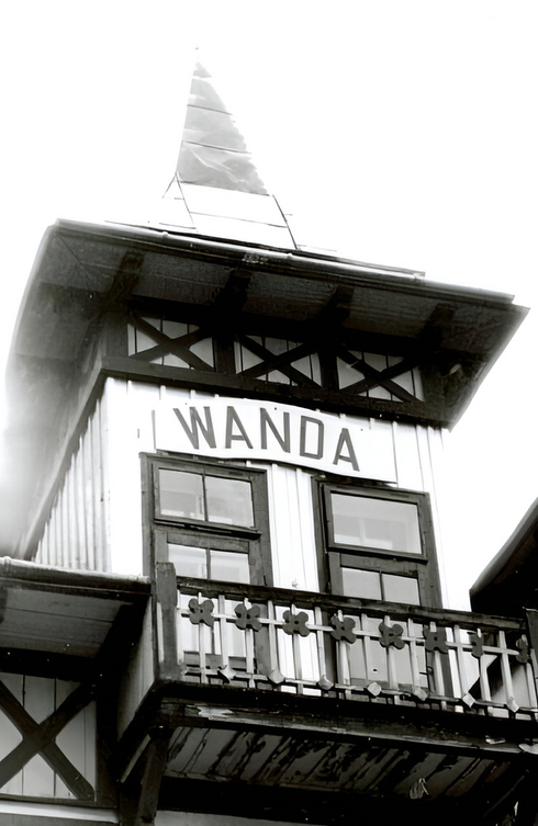 Fasada pensjonatu WANDA z wieżyczką w stylu muru pruskiego, ozdobnym drewnianym balkonem i małą chorągiewką na dachu, zabytkowy urok