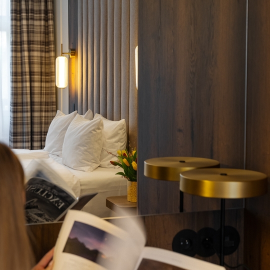 Przytulny pokój hotelowy z pikowanym zagłówkiem, białymi poduszkami, ciepłymi kinkietami, kraciastymi zasłonami i tulipanami; gość czyta magazyn.