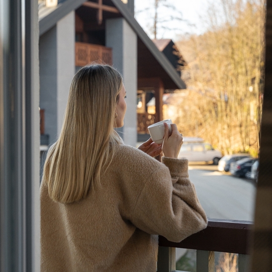 Gość na balkonie z filiżanką kawy w miękkim swetrze, widok na górski hotel w stylu chalet i słoneczny las.