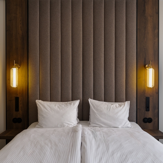 Dwa złączone łóżka z białą pościelą, wysoki tapicerowany zagłówek, ciepłe lampy nocne wiszące i drewniane panele