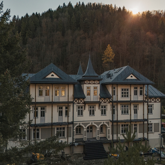 Zabytkowy górski pensjonat z trzema kondygnacjami, wieżyczką, łukowym wejściem i dużymi oknami na tle zalesionego stoku o zachodzie słońca.