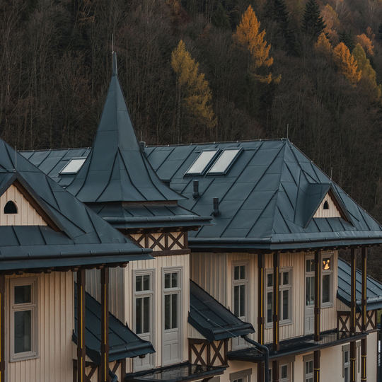 Fasada hotelu w alpejskim stylu z ciemnymi metalowymi dachami, wieżyczką, drewnianymi balkonami i oknami na tle jesiennego lasu.
