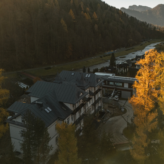 Górski hotel z ciemnymi metalowymi dachami przy wijącej się rzece, jesienne modrzewie w złotym świetle i widok na dolinę.