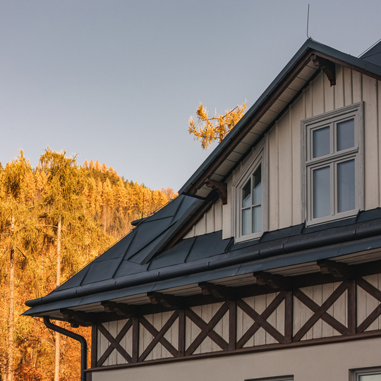 Szczyt górskiego pensjonatu z oknami mansardowymi, ciemnym metalowym dachem i drewnianymi belkami, złoty jesienny las w tle.