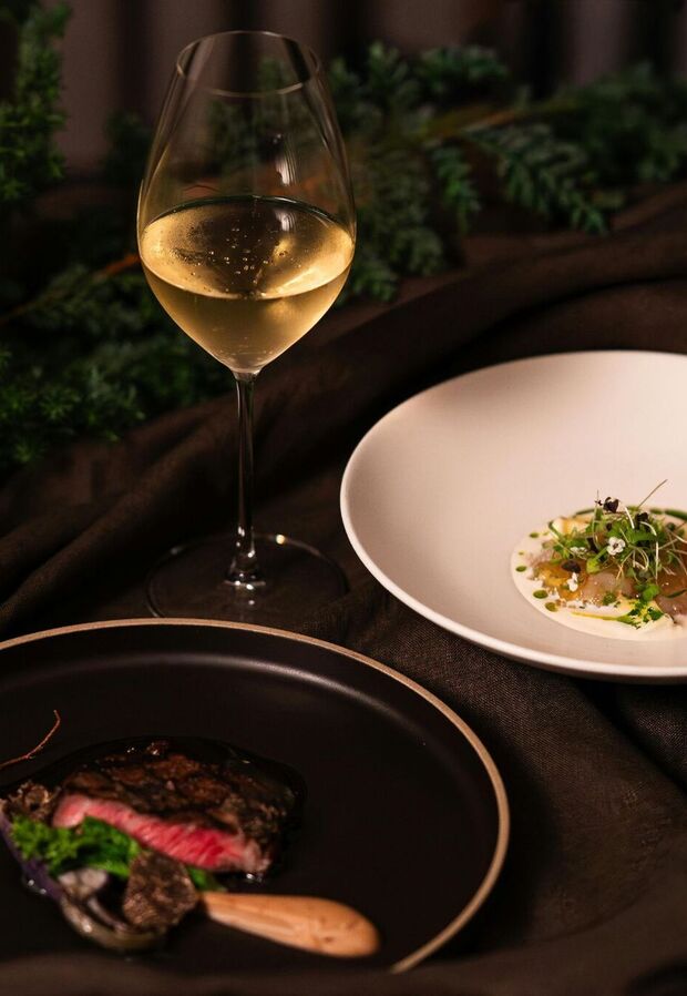 Kieliszek schłodzonego białego wina obok steku na czarnej porcelanie i delikatnej przystawki z mikroliśćmi, nastrojowa aranżacja stołu