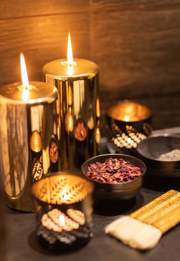 Palące złote świece słupkowe i ażurowe lampiony obok miseczki z suszonymi płatkami róż i pałeczek zapachowych, ciepła atmosfera spa.