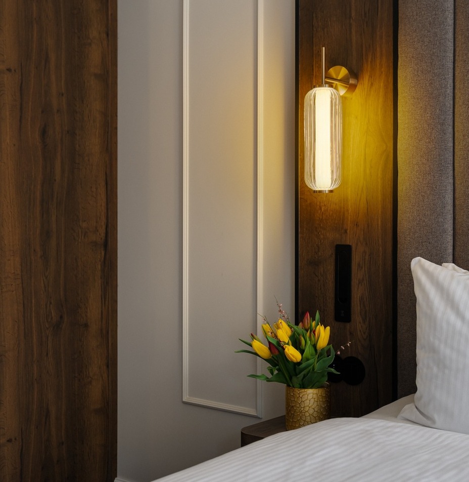 Ciepły kinkiet nad drewnianym zagłówkiem, śnieżnobiała pościel i złoty wazon z żółtymi tulipanami na stoliku.