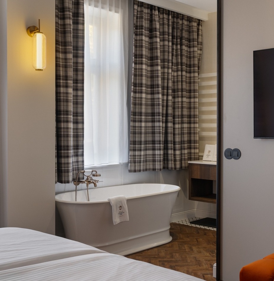 Wolnostojąca biała wanna przy oknie z zasłonami w kratę, ciepły kinkiet, telewizor na ścianie i pomarańczowy puf.